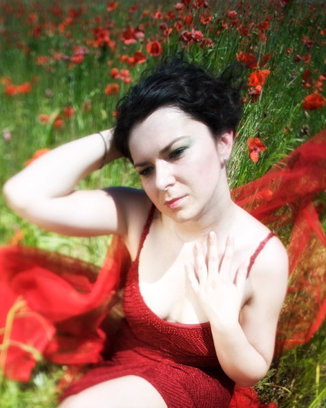 Kobieta w czerwonej sukience pozuje w plenerze na łące z makami, dłoń na dekolcie, wzrok skierowany w dół.