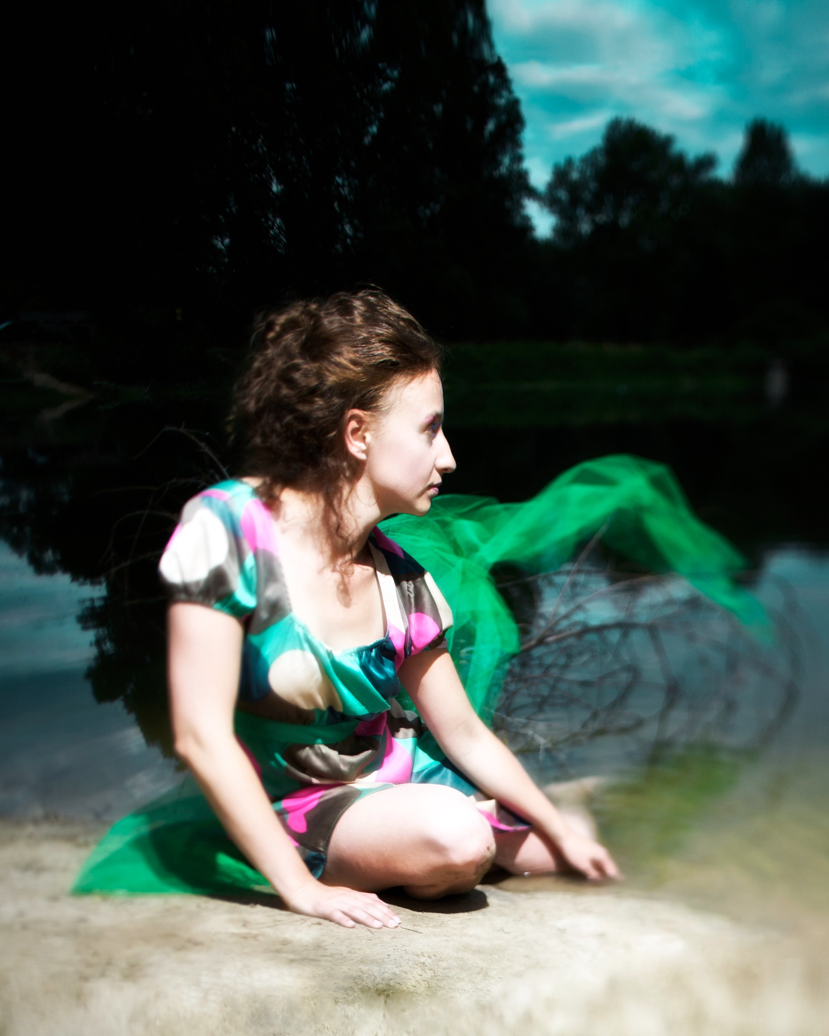 Młoda kobieta w sukience w plenerze, siedząca na brzegu rzeki z zieloną tkaniną, profil, zamyślony wyraz twarzy, ciemne tło z drzewami.