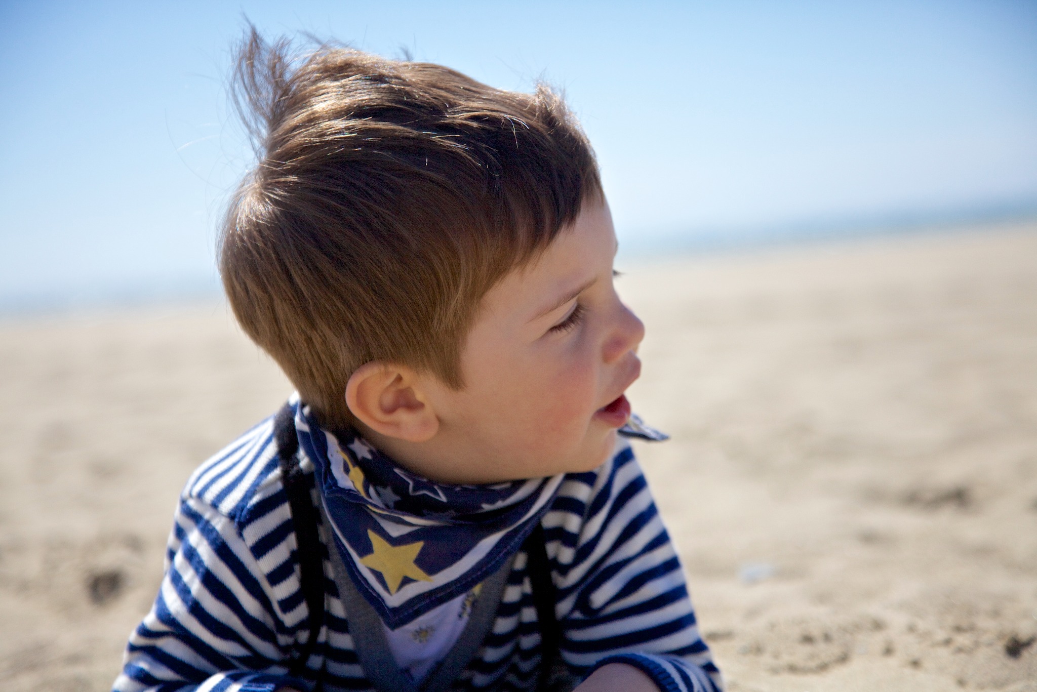 Portret z profilu małego chłopca na plaży, ubrany w bluzkę w paski i chustę w gwiazdki, z rozwichrzonymi włosami na tle piasku i błękitnego nieba.