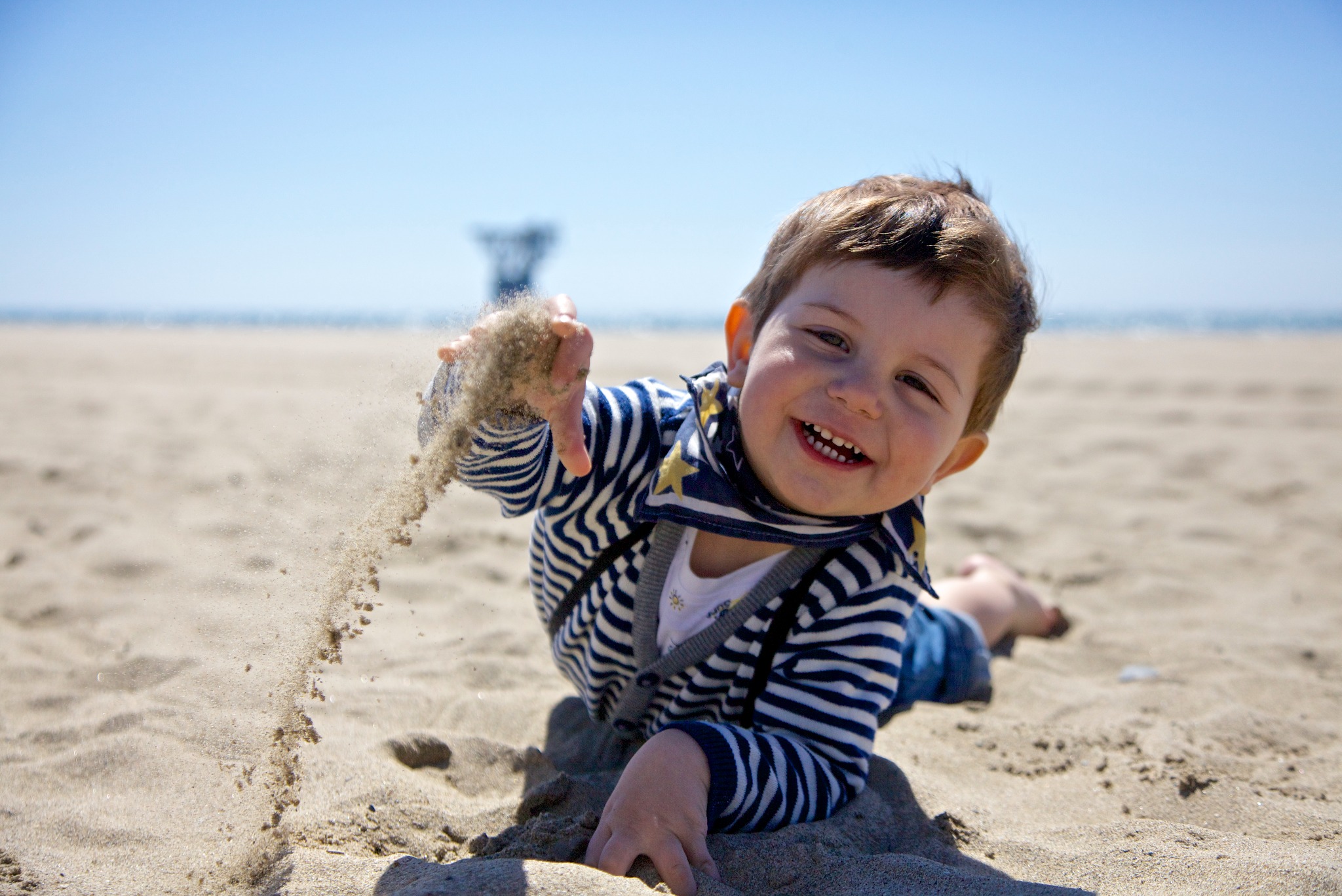 Uśmiechnięty chłopiec w pasiastym swetrze leży na piaszczystej plaży w Luboniu, bawiąc się piaskiem, który przesypuje się przez jego dłoń na tle błękitnego nieba i morza.