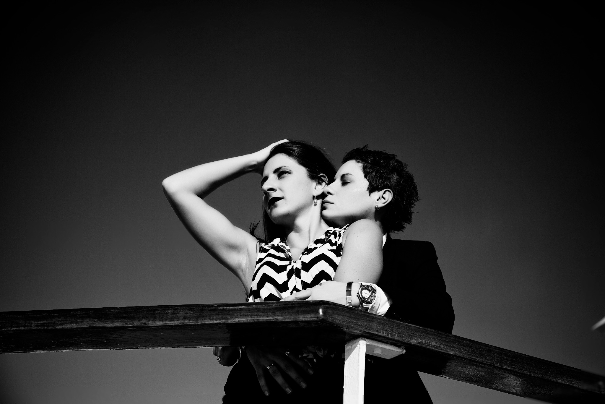 Czarno-białe zdjęcie portretowe dwóch kobiet w plenerze, jedna w sukience w zygzaki, druga w marynarce, opierają się o drewnianą barierkę, z głowami blisko siebie.