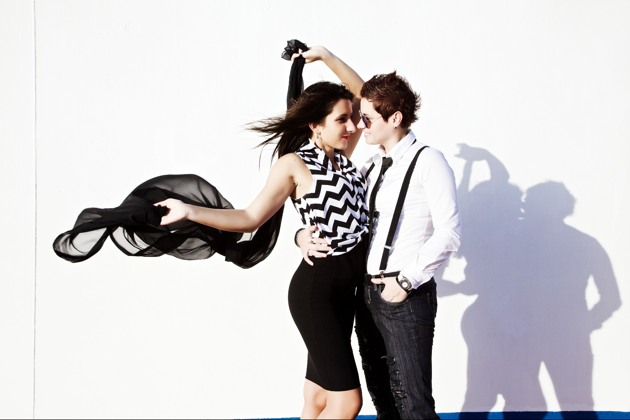 Para w stylizowanych ubraniach na tle białej ściany: kobieta w czarno-białej bluzce i czarnej spódnicy, trzymająca czarną, zwiewną chustę, mężczyzna w białej koszuli z szelkami i ciemnych jeansach,...