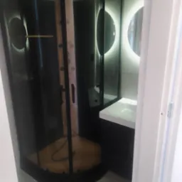 Nowoczesna kabina prysznicowa z czarnymi profilami i drewnianym panelem wewnątrz, okrągłe lustro z podświetleniem LED, minimalistyczna umywalka