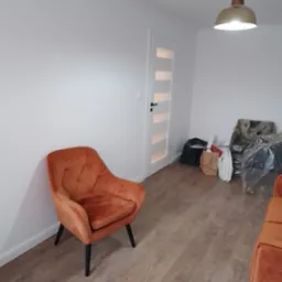 Pokój po remoncie z pomarańczowym fotelem, drewnopodobną podłogą i białymi ścianami; widoczne worki i kartony po meblach w tle.