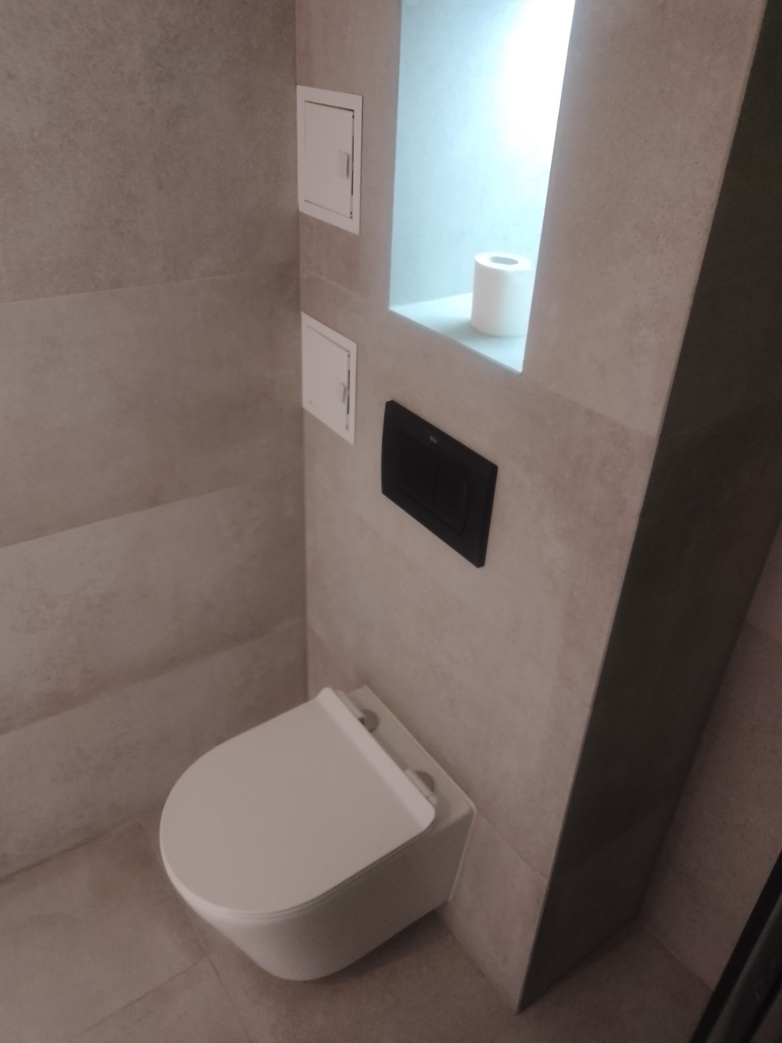 Nowoczesna toaleta podwieszana z czarnym przyciskiem spłukującym na tle beżowych płytek ceramicznych. W ścianie wnęka z rolką papieru toaletowego.