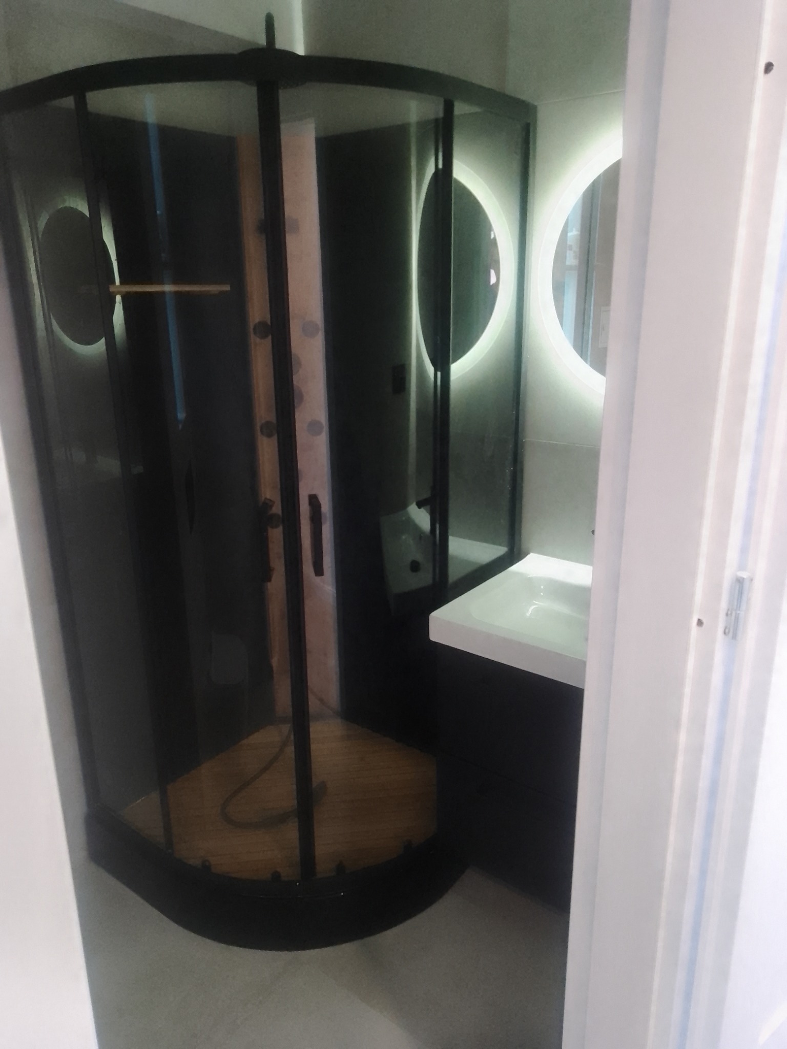 Nowoczesna kabina prysznicowa z czarnymi profilami i drewnianym panelem wewnątrz, okrągłe lustro z podświetleniem LED, minimalistyczna umywalka