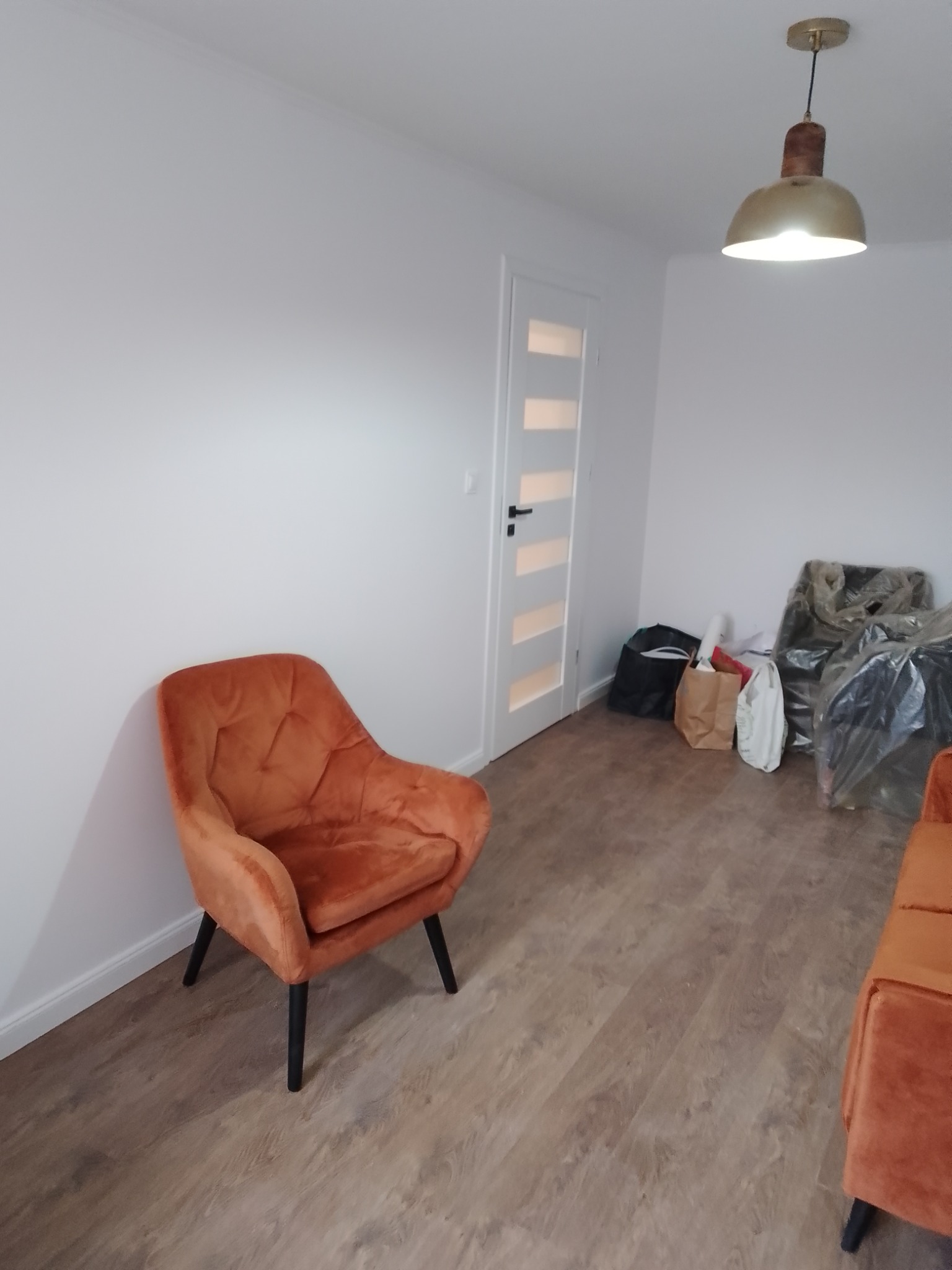 Pokój po remoncie z pomarańczowym fotelem, drewnopodobną podłogą i białymi ścianami; widoczne worki i kartony po meblach w tle.