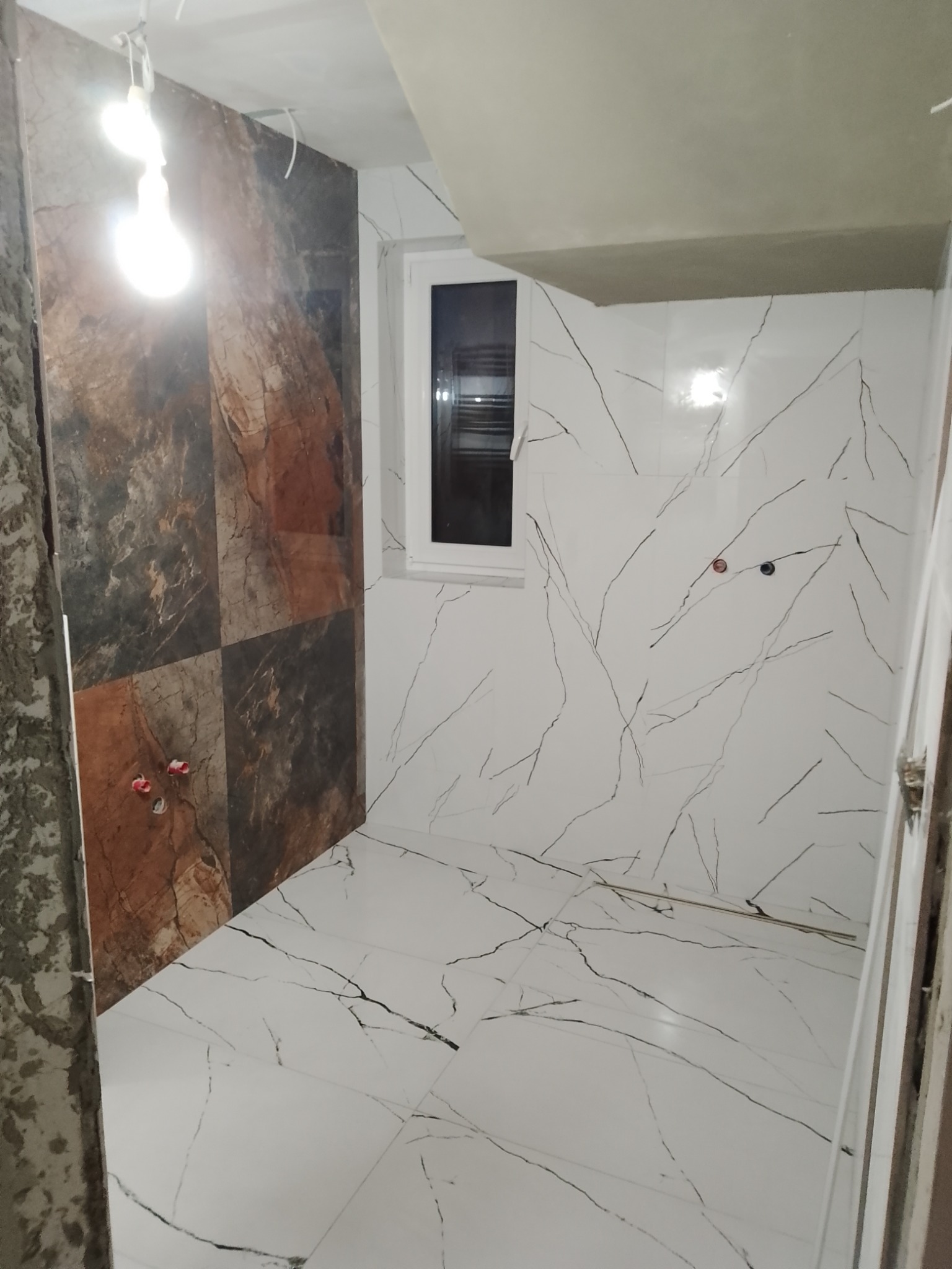 Wnętrze łazienki w trakcie remontu, z częściowo położonymi płytkami imitującymi marmur na podłodze i ścianach, widoczne wystające rury i surowy sufit.