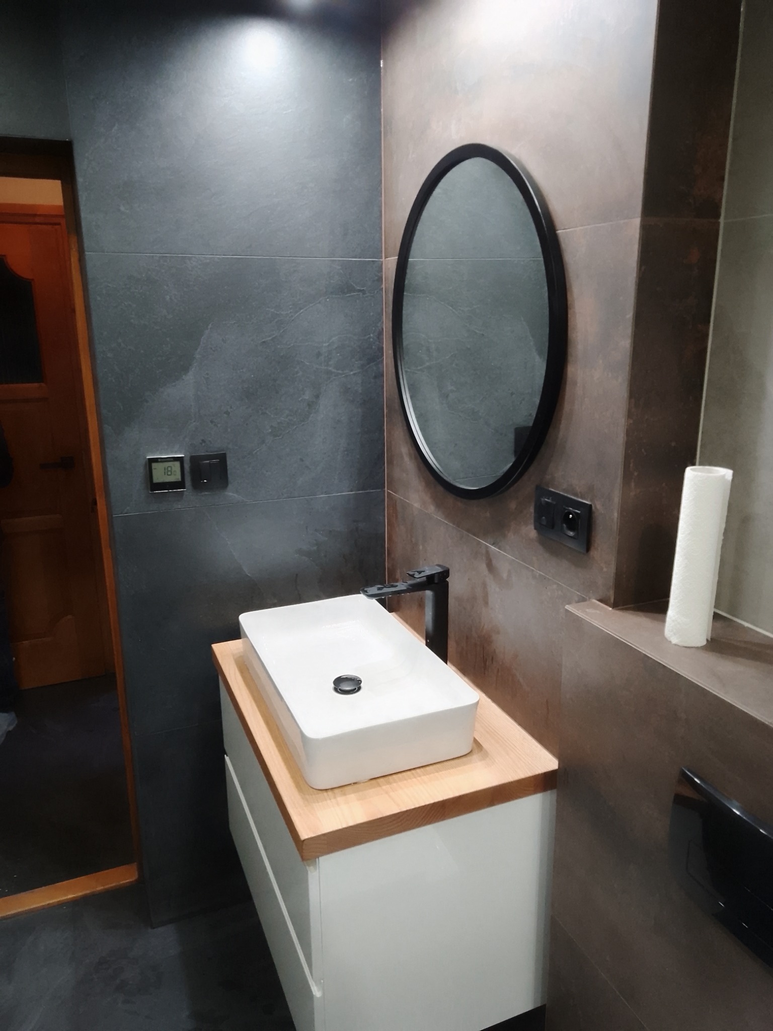 Nowoczesna łazienka z ciemnoszarymi i brązowymi płytkami, owalnym lustrem w czarnej ramie nad białą szafką z drewnianym blatem i prostokątną umywalką.