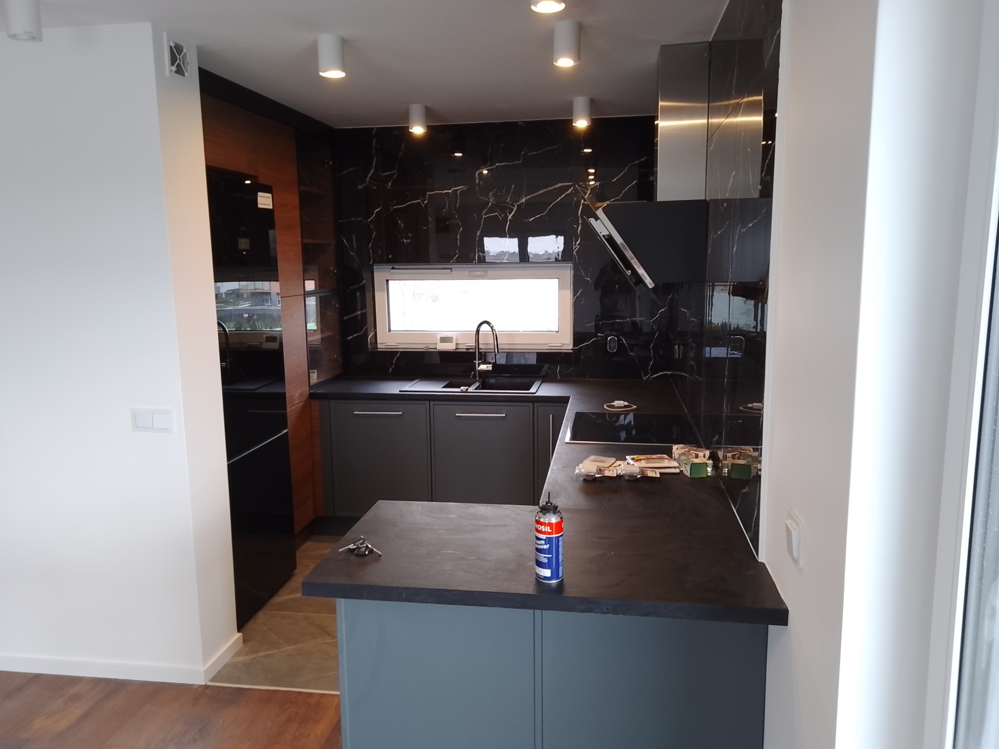 Nowoczesna kuchnia z czarnymi marmurkowymi płytkami na ścianie, szarymi szafkami i blatem, widoczna puszka z pianką montażową na wyspie kuchennej, klucze leżące obok.