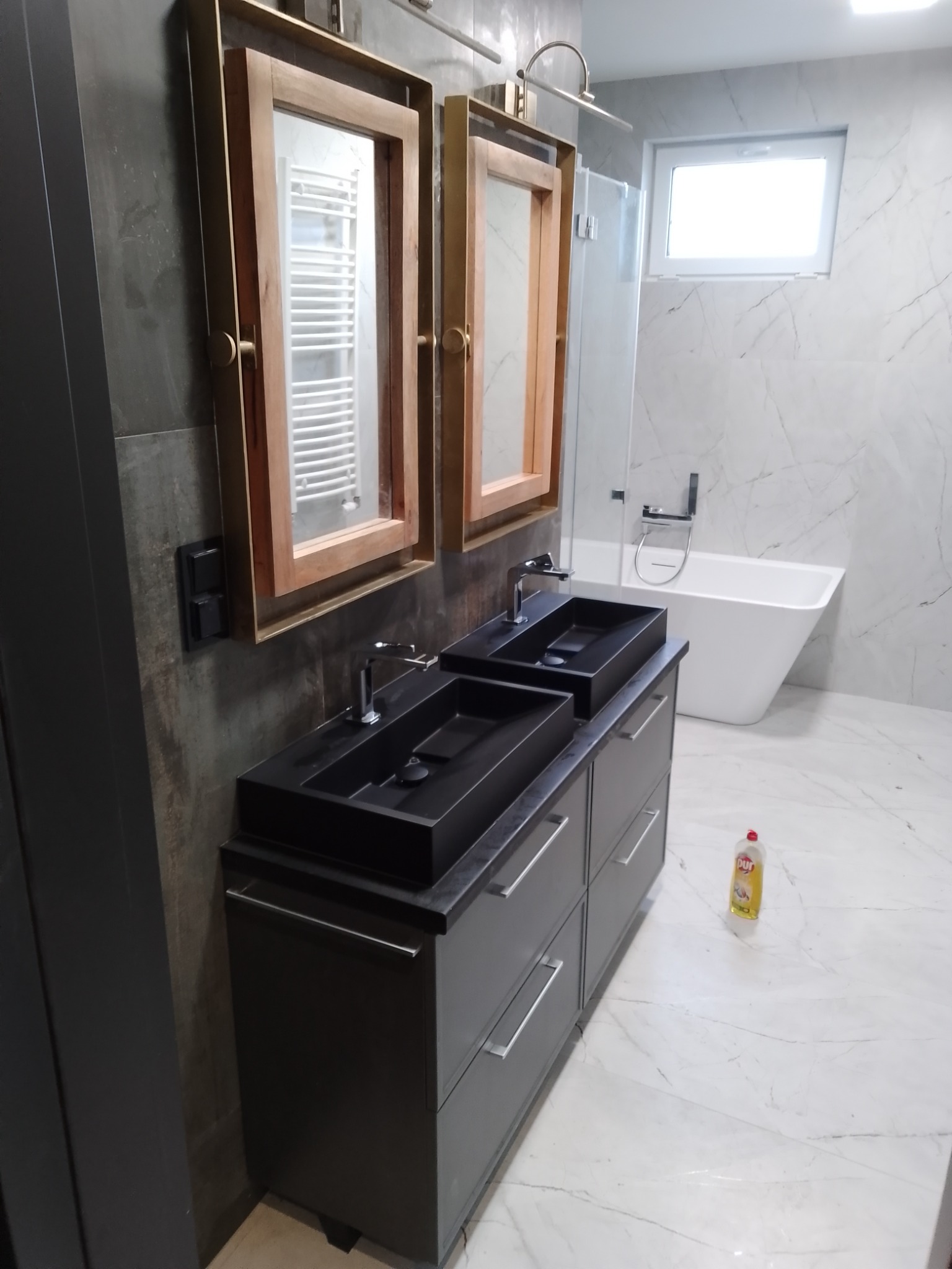 Nowoczesna łazienka z podwójną czarną umywalką nablatową na szarej szafce, lustra w drewnianych ramach i biała wanna wolnostojąca na tle marmurowych płytek. Na podłodze stoi butelka płynu do mycia.