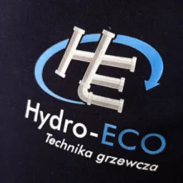 Granatowa koszulka z nadrukowanym logo firmy Hydro-ECO Technika grzewcza, przedstawiającym stylizowane rury w kształcie liter HE otoczone niebieską strzałką.