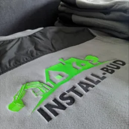 Szara bluza polarowa z wyhaftowanym zielonym logo firmy budowlanej 'INSTALL-BUD', obok leżą szare rękawice robocze.