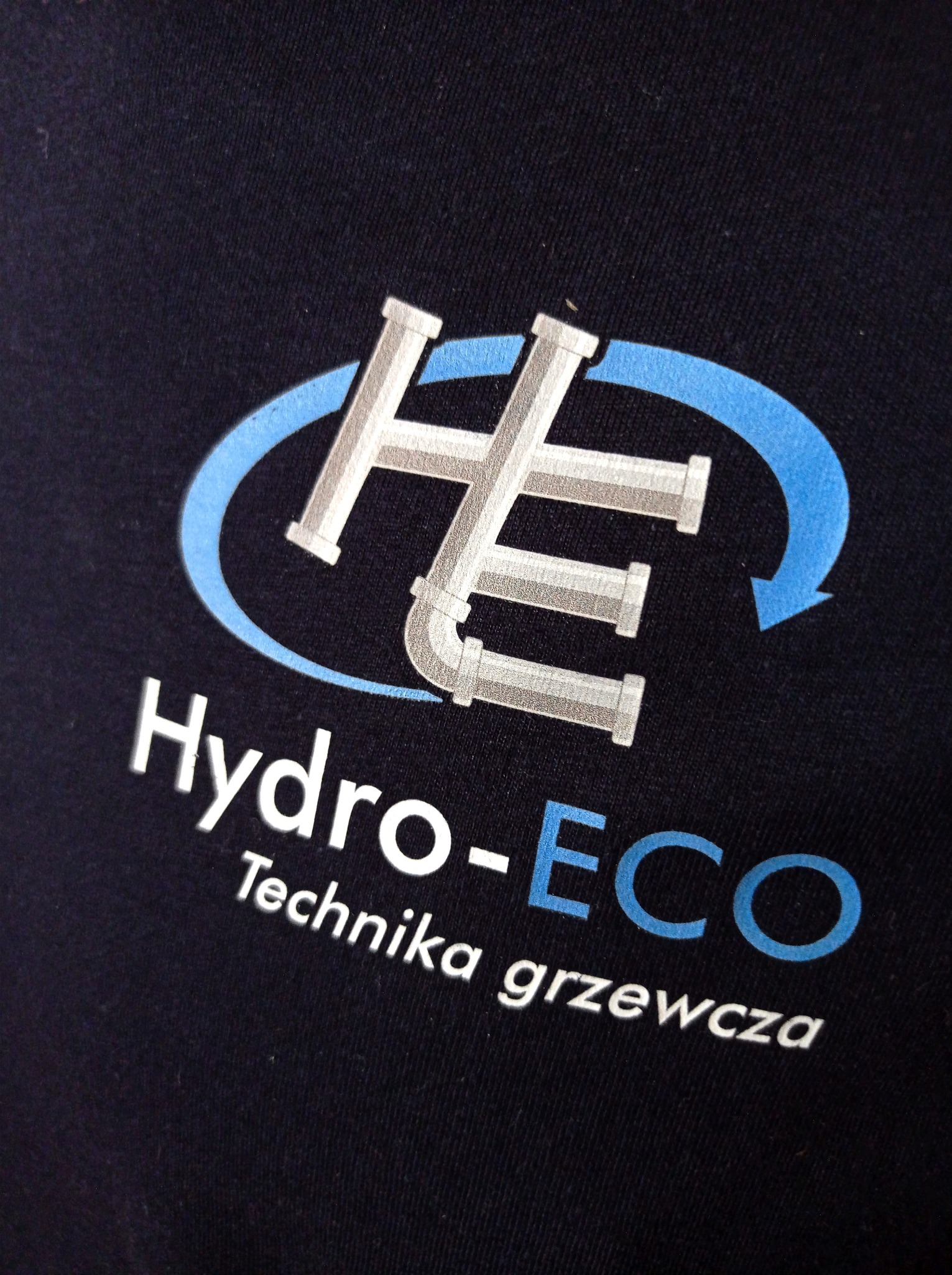 Granatowa koszulka z nadrukowanym logo firmy Hydro-ECO Technika grzewcza, przedstawiającym stylizowane rury w kształcie liter HE otoczone niebieską strzałką.