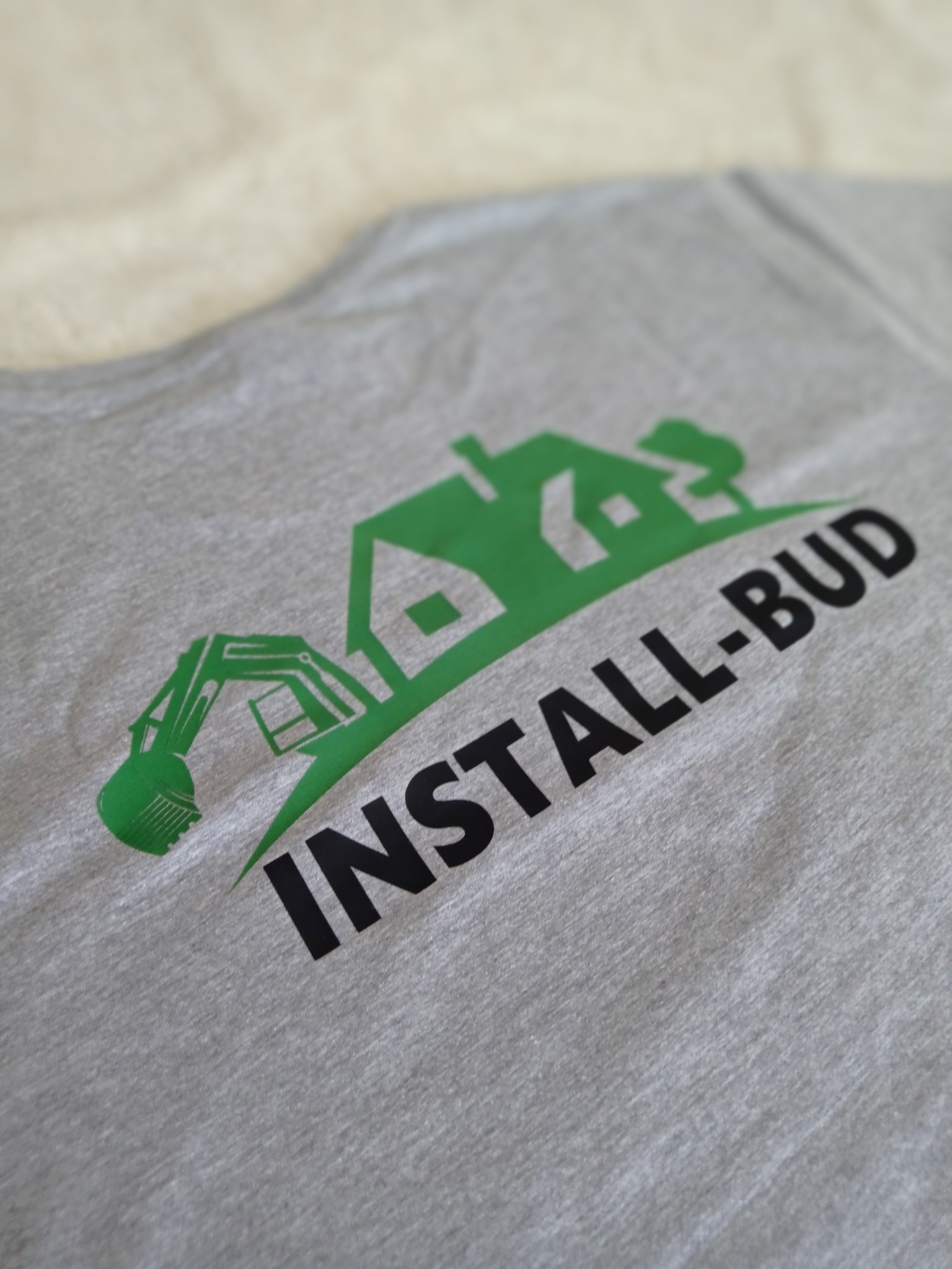 Szara koszulka z zielonym nadrukiem logo firmy budowlanej przedstawiającym dom, drzewo i koparkę, z czarnym napisem 'INSTALL-BUD'.