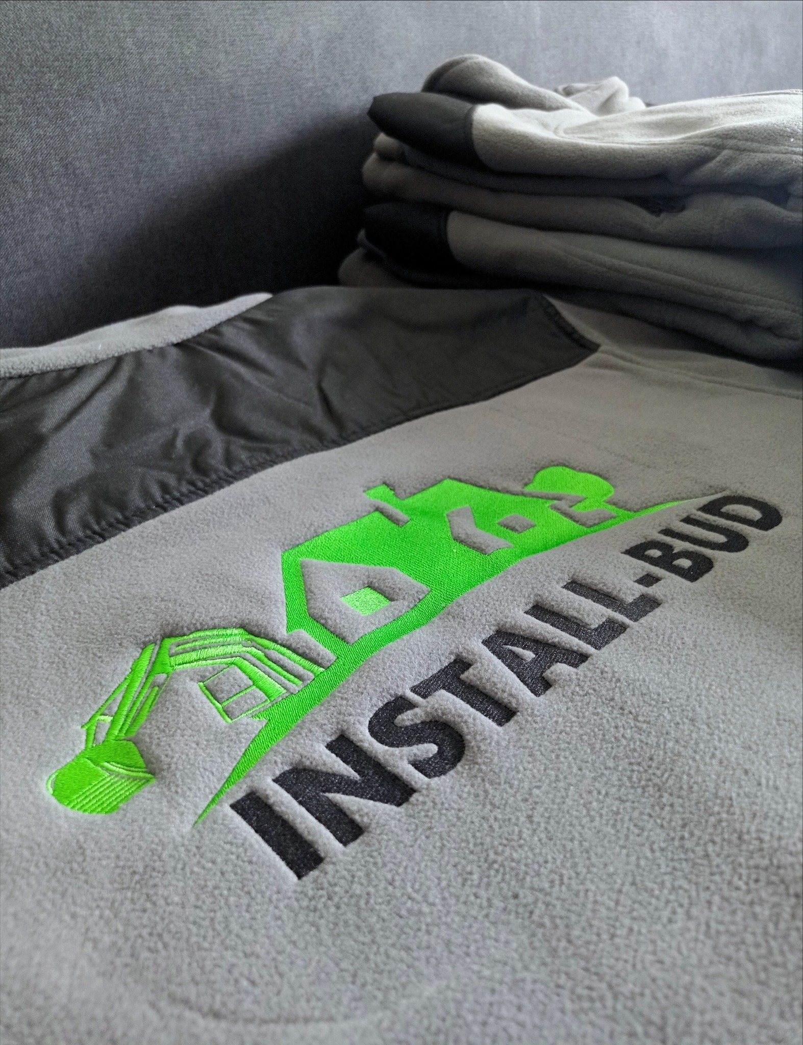 Szara bluza polarowa z wyhaftowanym zielonym logo firmy budowlanej 'INSTALL-BUD', obok leżą szare rękawice robocze.