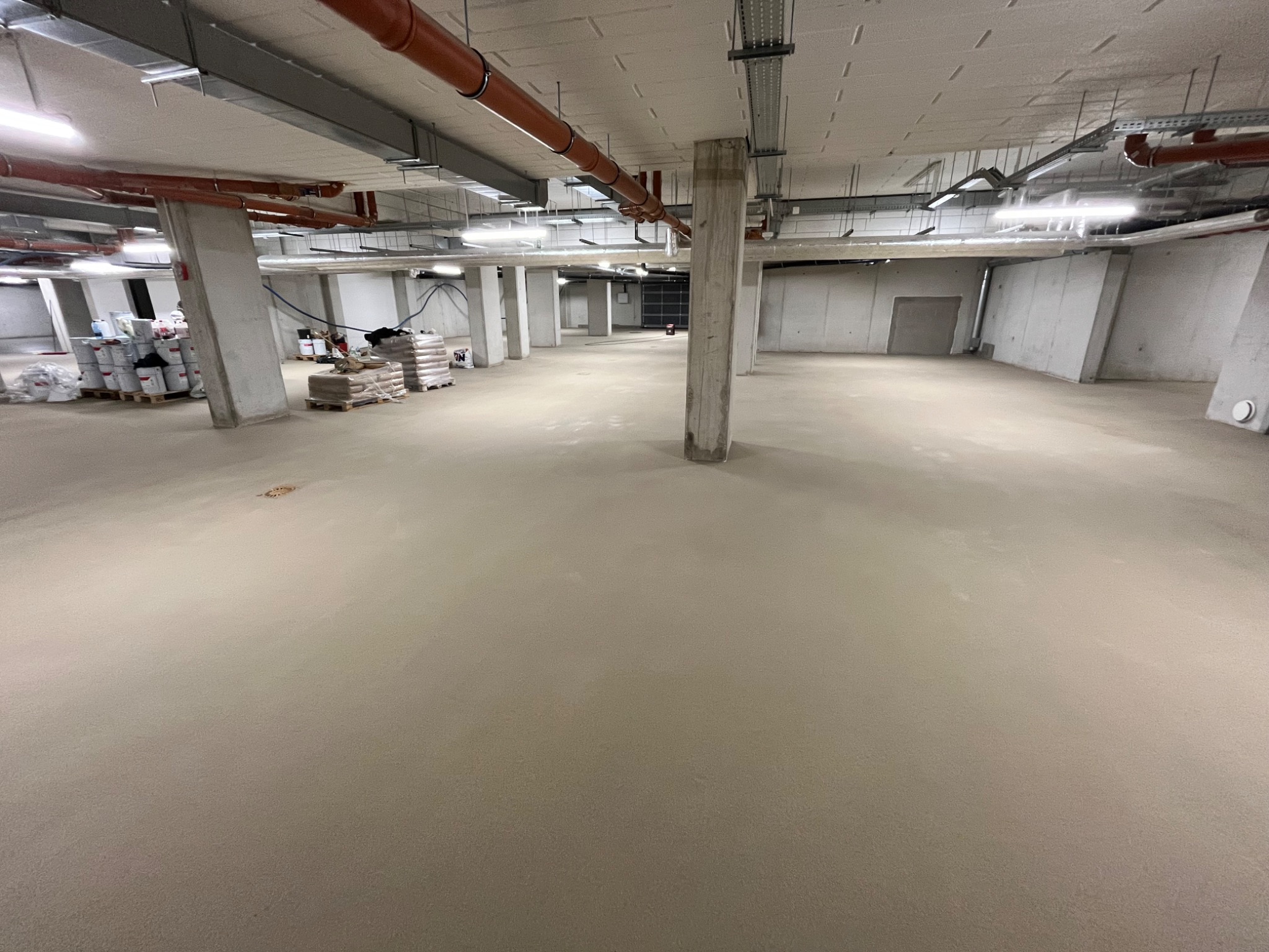 Wykończona, jasnoszara, gładka podłoga w dużej, pustej hali przemysłowej z betonowymi kolumnami, widocznymi instalacjami pod sufitem i paletami z materiałami budowlanymi w tle.