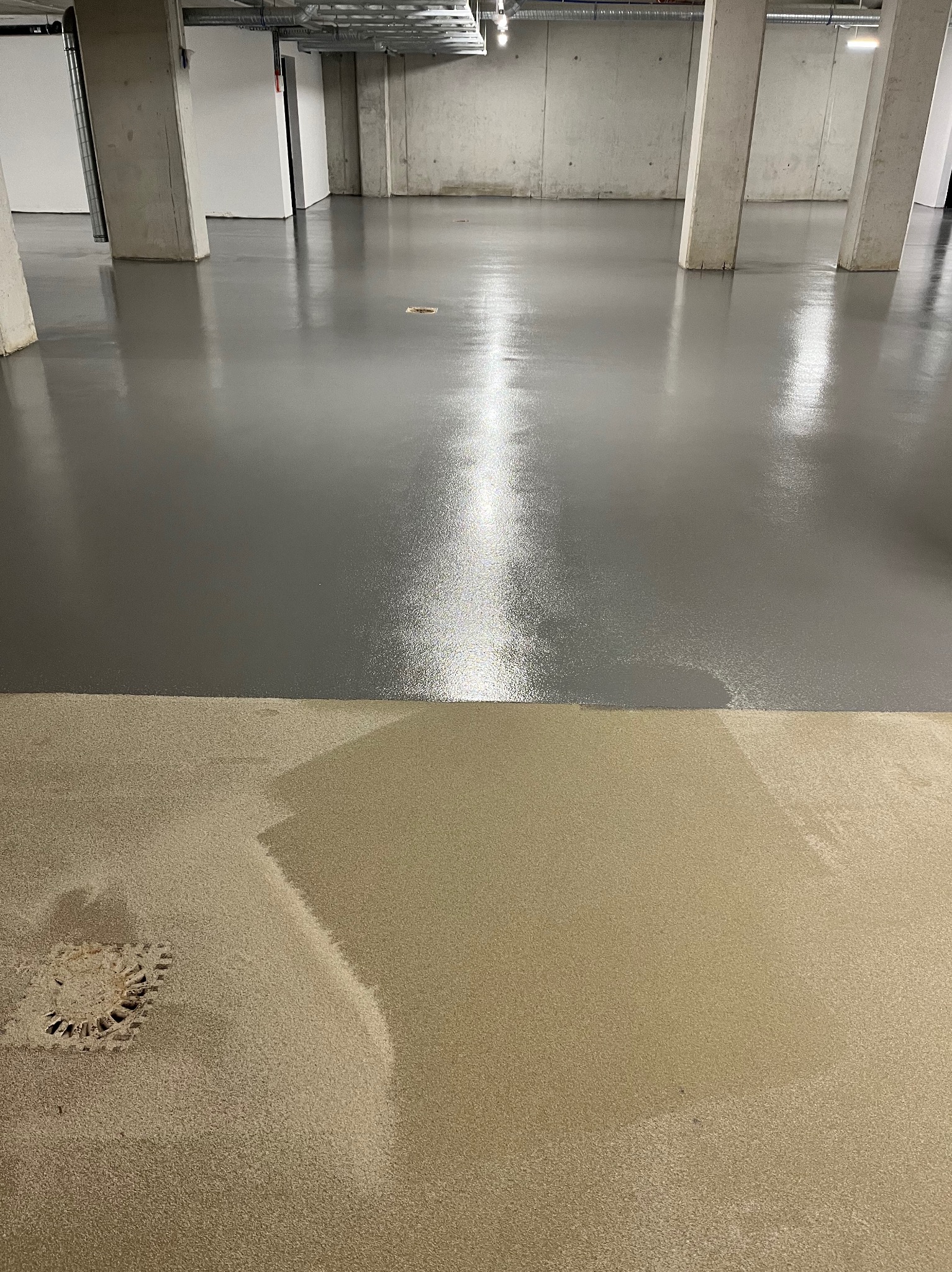 Szara, błyszcząca podłoga w hali przemysłowej, częściowo pokryta beżową warstwą, widoczne słupy konstrukcyjne i studzienka kanalizacyjna w dolnej części kadru.