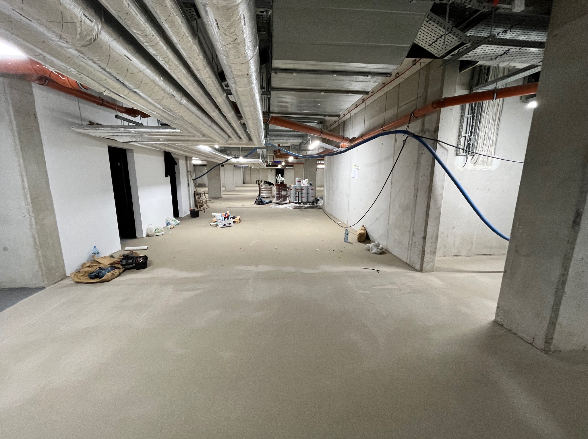 Jasna, świeżo wylana posadzka przemysłowa w surowym wnętrzu budynku, z widocznymi instalacjami pod sufitem i roboczym bałaganem w tle.