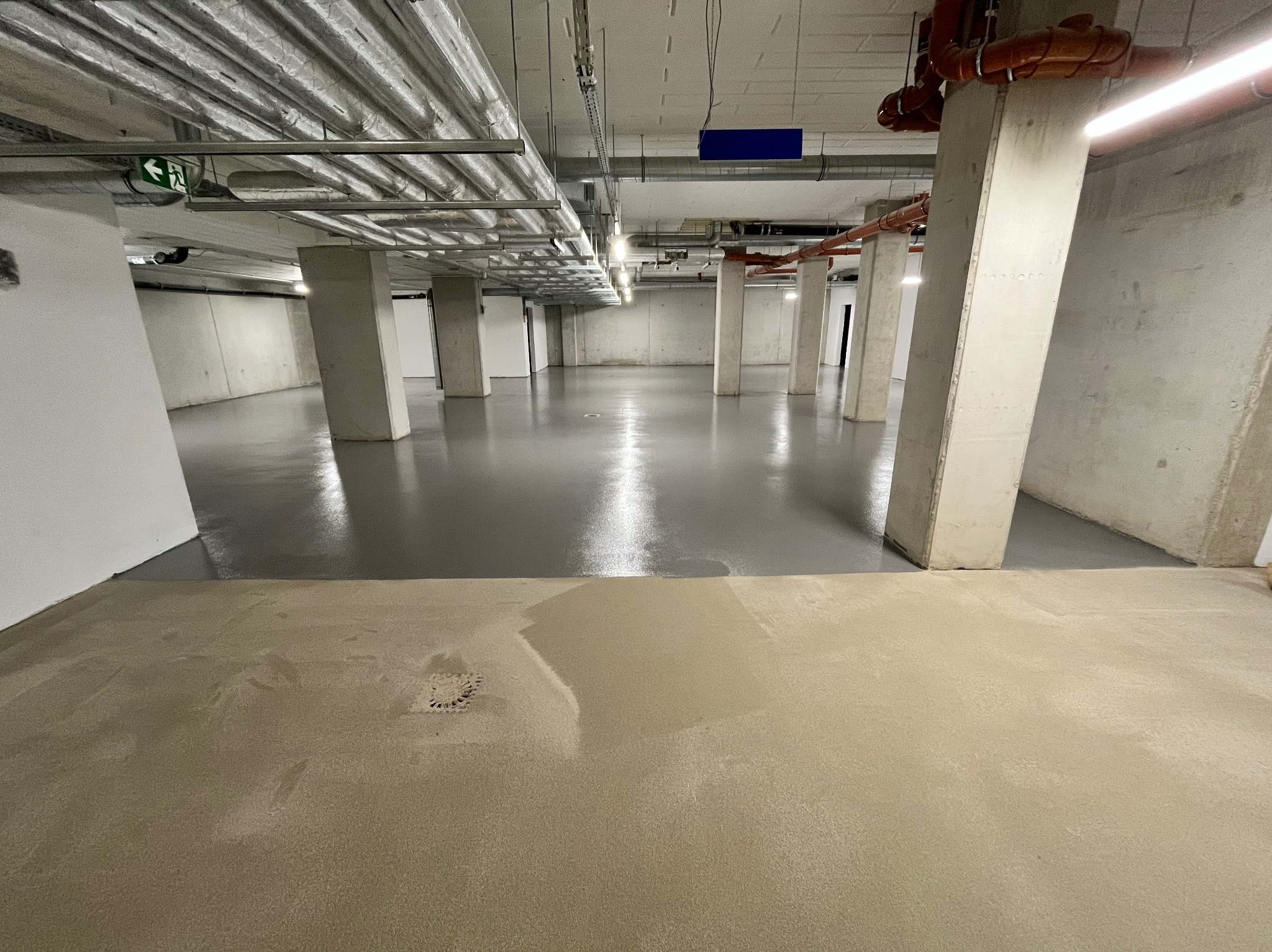 Wykończona szara podłoga w hali przemysłowej z widocznymi betonowymi kolumnami, rurami pod sufitem i kratką ściekową w dolnym rogu zdjęcia, część podłogi jeszcze nie wykończona.