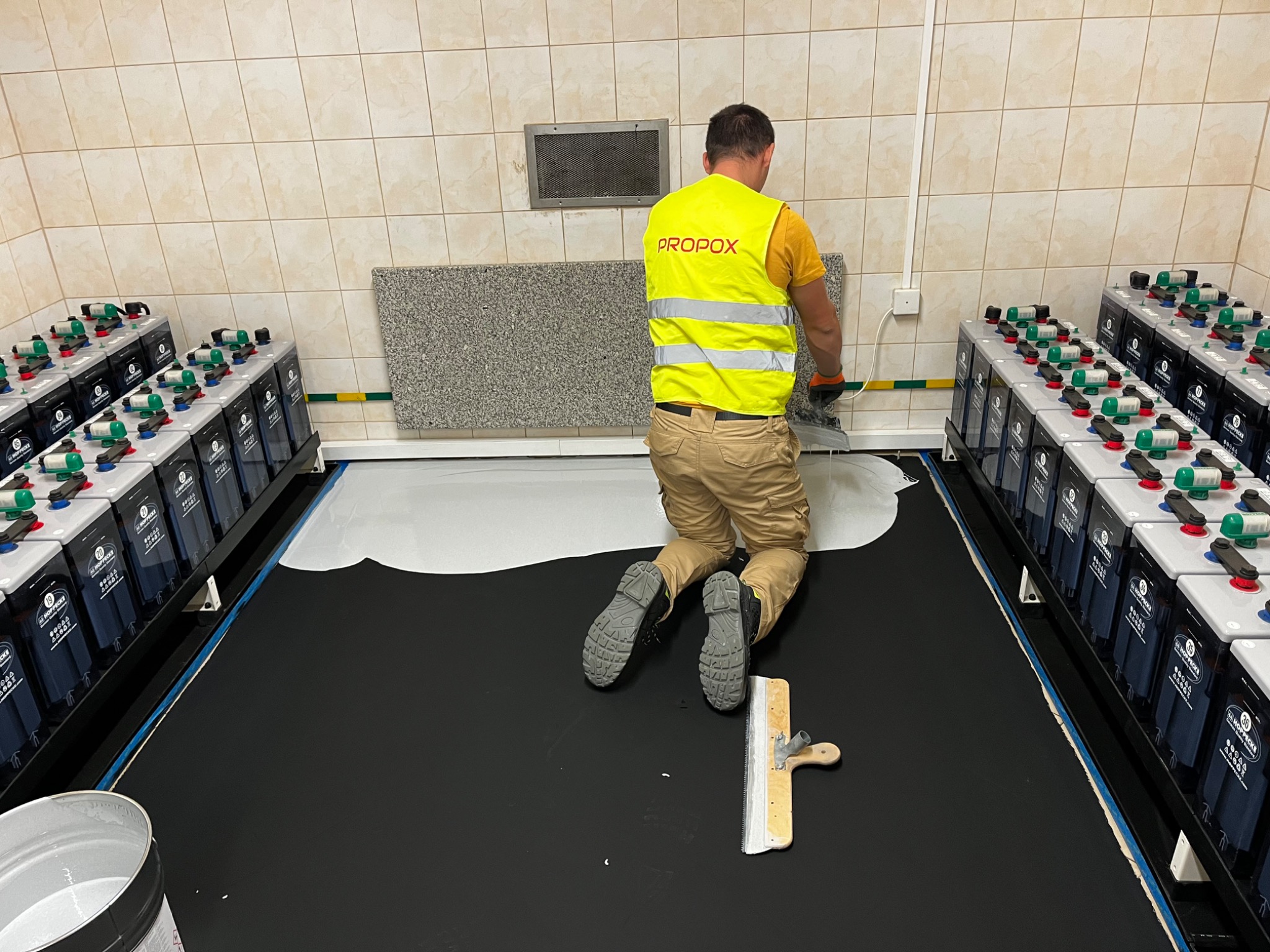 Pracownik w żółtej kamizelce z logo PROPOX rozprowadza białą substancję na czarnej podłodze w pomieszczeniu z rzędami baterii po bokach i kafelkowanymi ścianami.