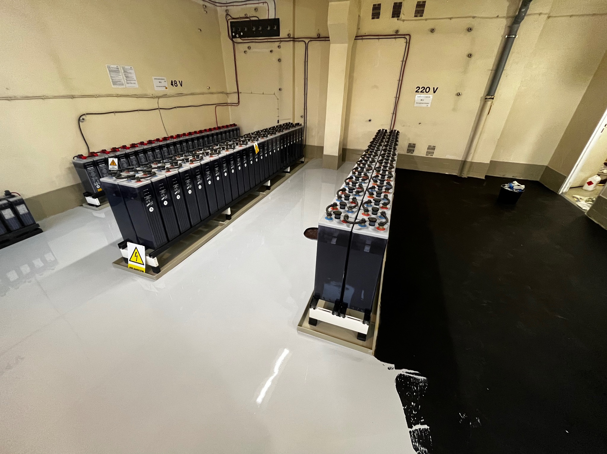 Pomieszczenie z dwiema rzędami akumulatorów przemysłowych na posadzce żywicznej, częściowo czarnej, częściowo białej, widoczne okablowanie 48V i 220V na ścianach.