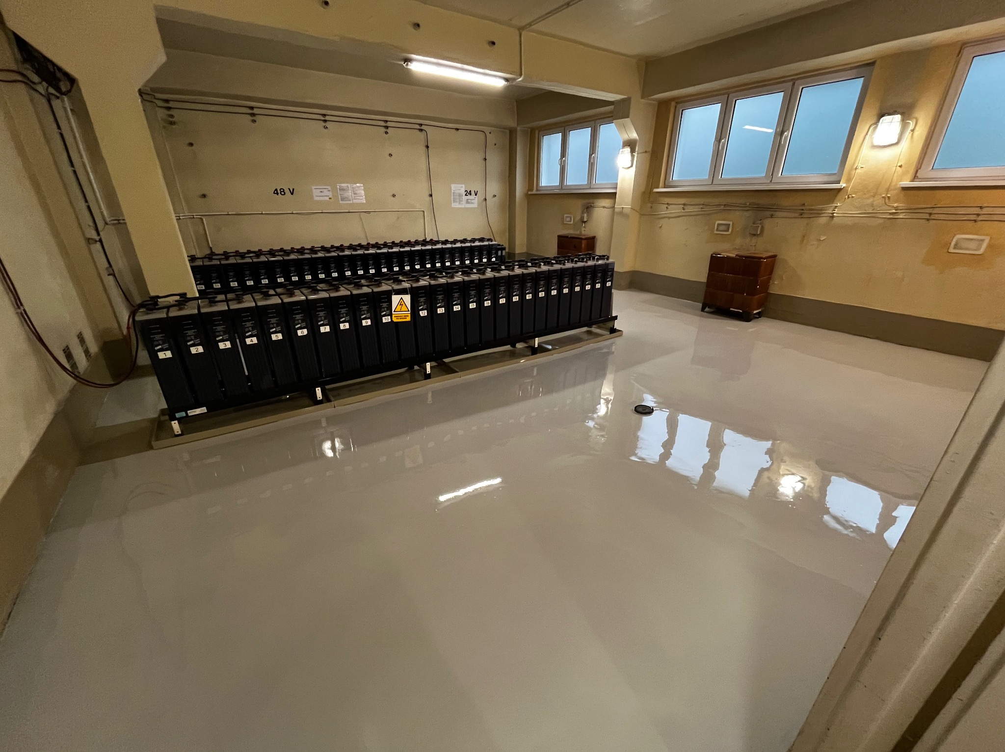 Jasnoszara, gładka posadzka w pomieszczeniu technicznym z rzędami baterii, widoczne okna i instalacje elektryczne na ścianach.