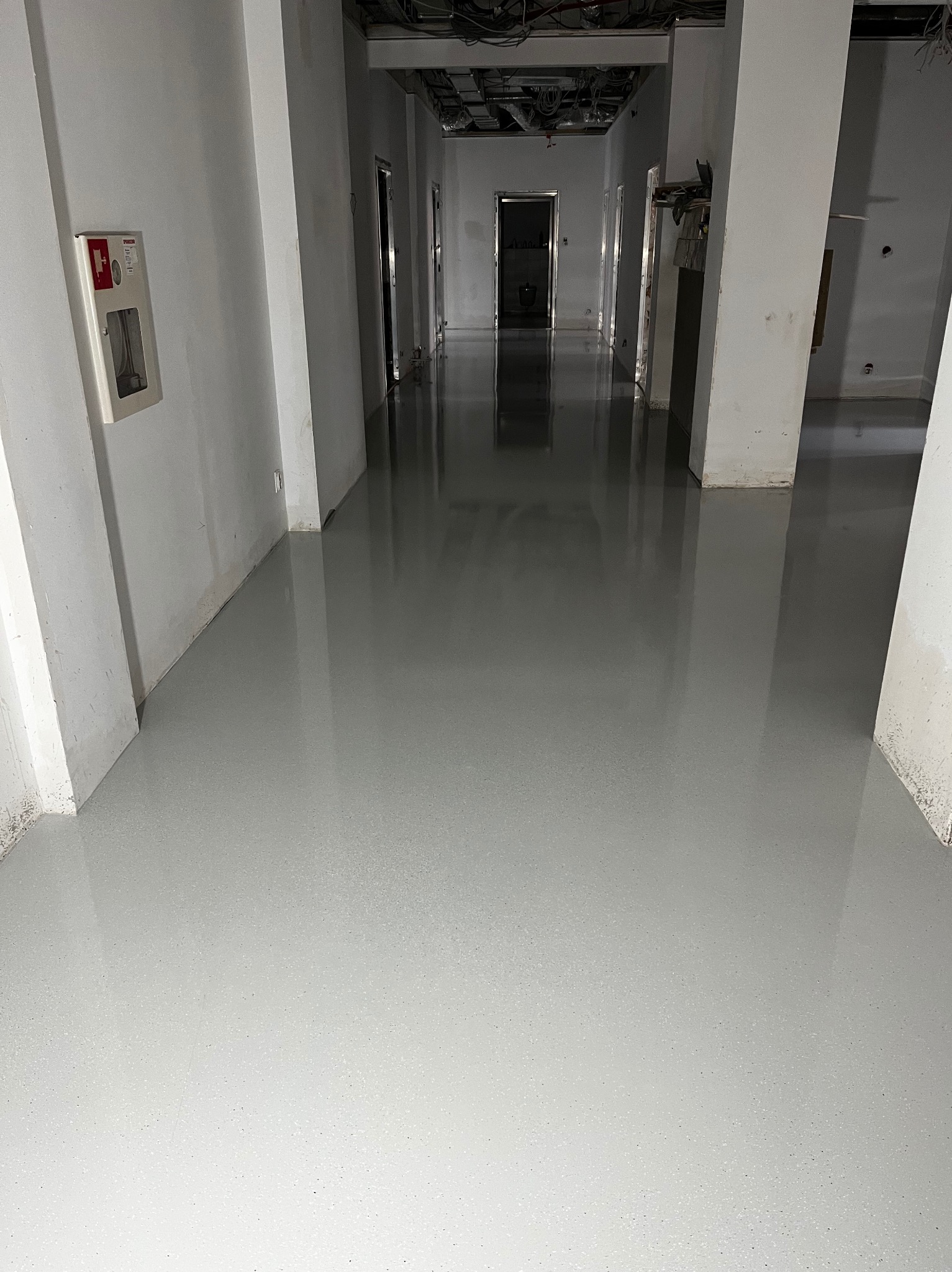Długi korytarz z świeżo wylaną, gładką, szarą posadzką. Widoczne filary i instalacje pod sufitem w stanie surowym. Na ścianie skrzynka przeciwpożarowa.