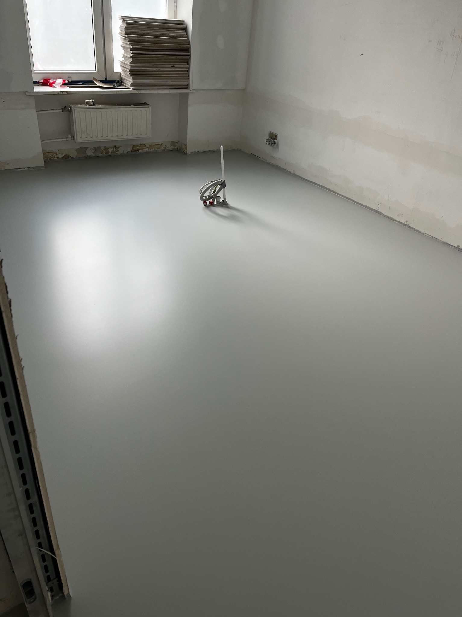 Świeżo wylana, szara, gładka podłoga w trakcie realizacji, z widocznym narzędziem do mieszania farb na środku pomieszczenia i stertą płyt na parapecie okna.