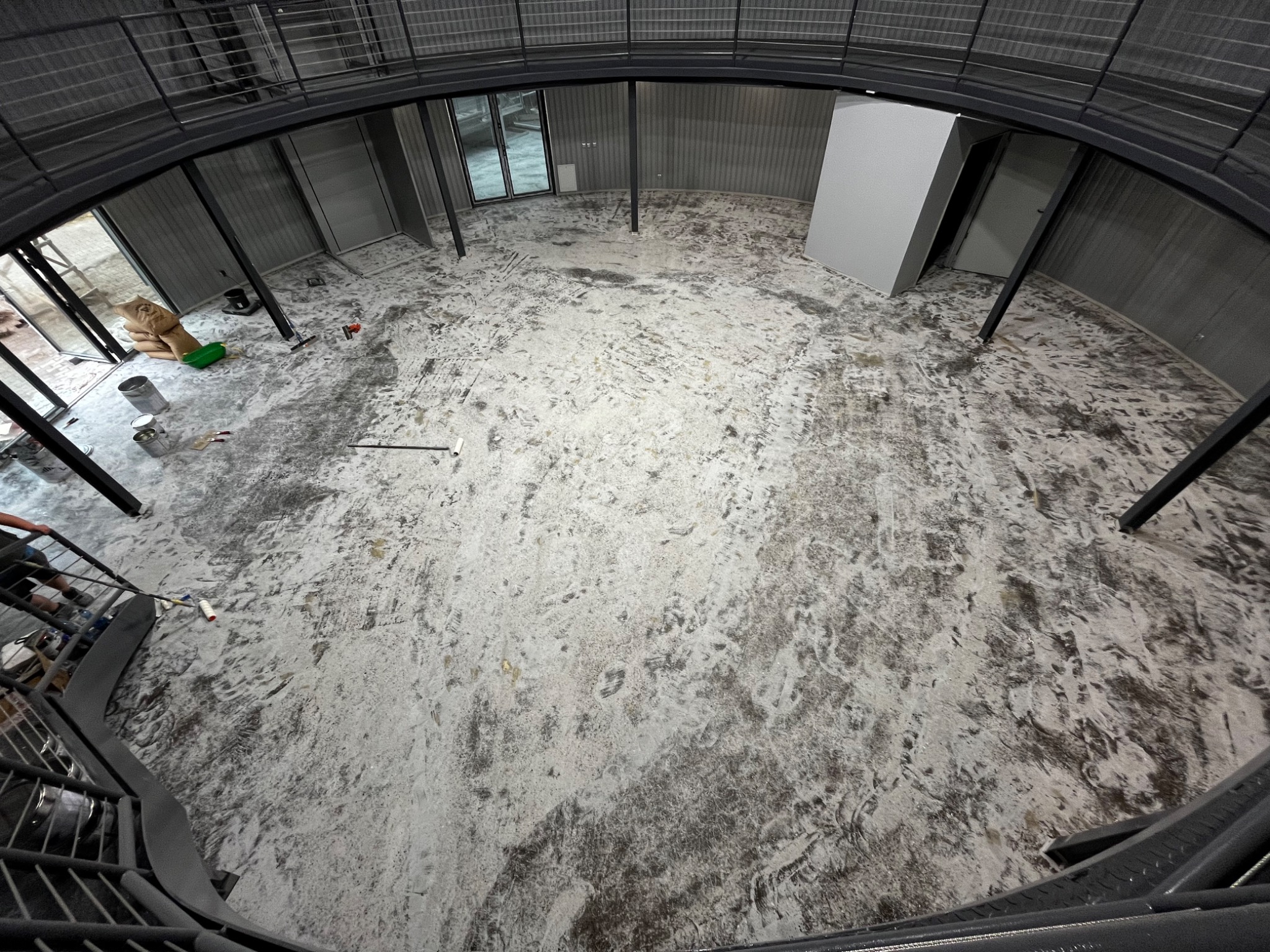 Wnętrze okrągłego budynku w Piławie Górnej w trakcie prac wykończeniowych posadzki, widoczne narzędzia i materiały budowlane oraz ślady stóp na surowej powierzchni.
