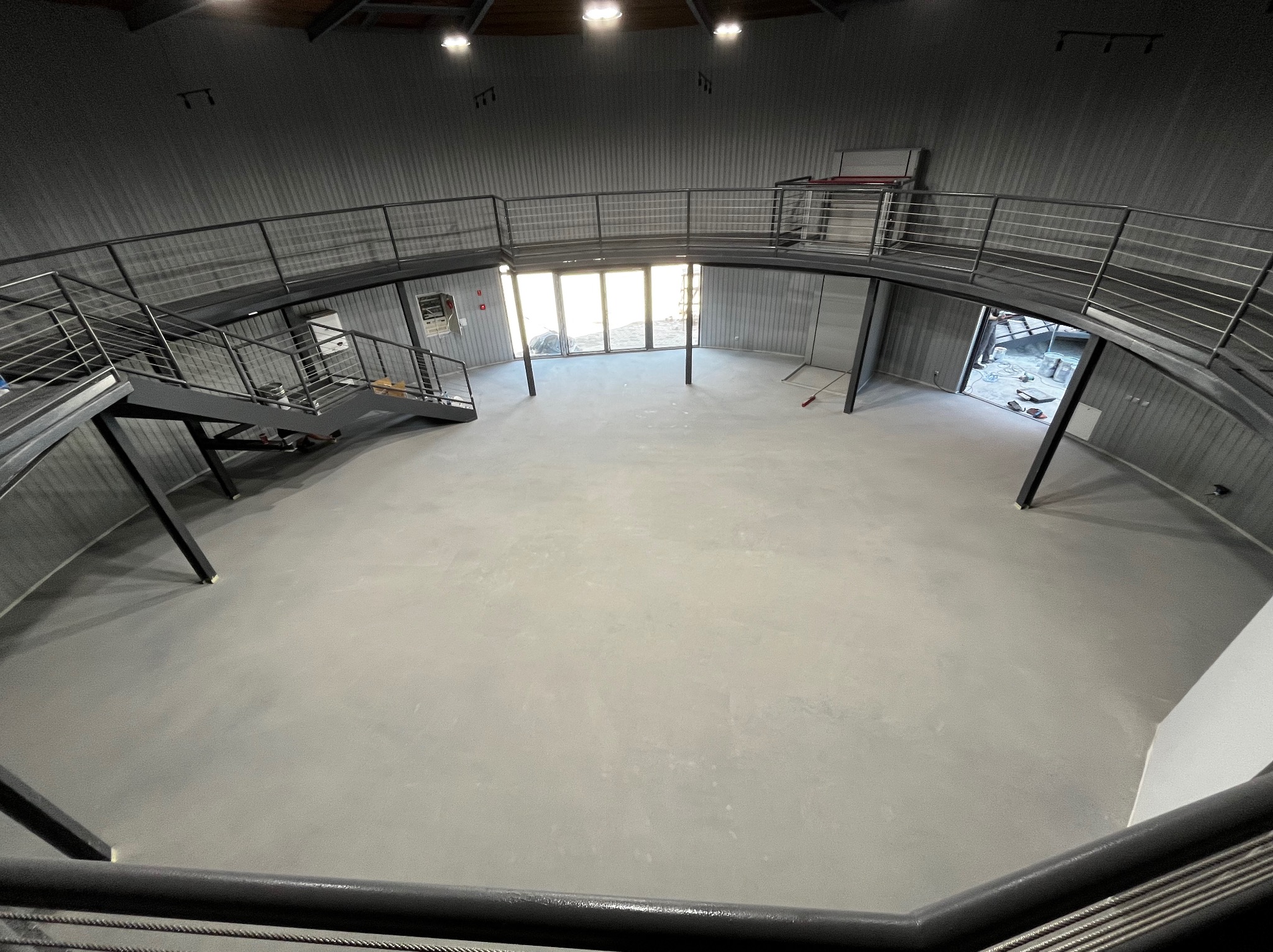 Wnętrze hali przemysłowej z szarą, gładką posadzką, widok z antresoli z metalową balustradą, w tle widoczne przeszklone drzwi i wejście do kolejnego pomieszczenia.