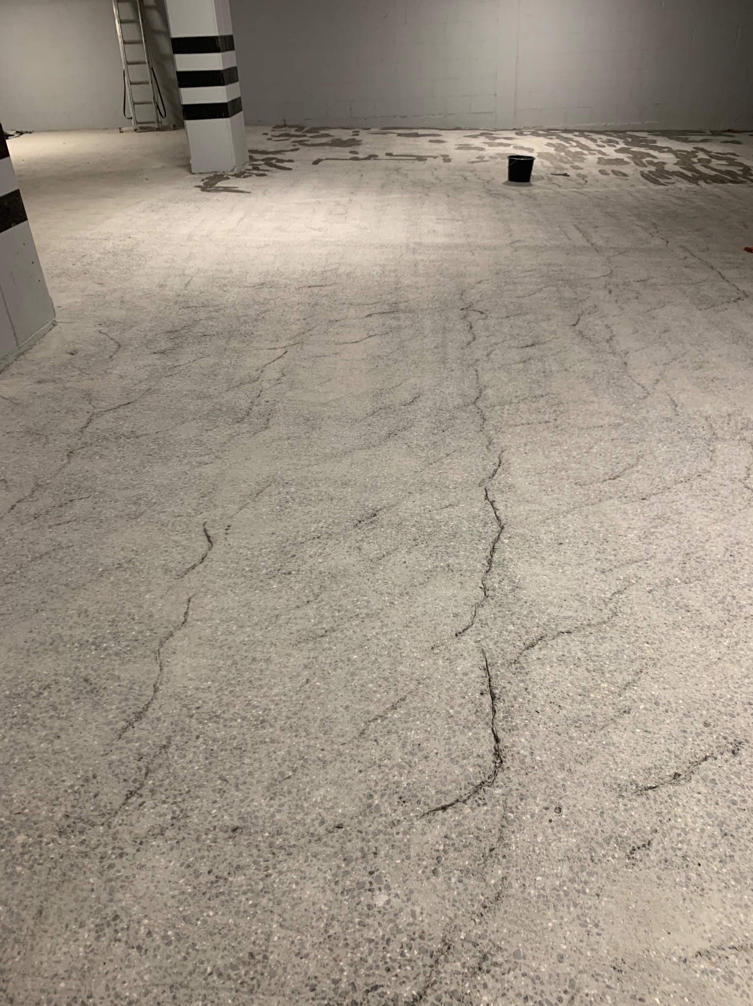 Szara, popękana, betonowa podłoga w hali przemysłowej, z czarnym wiadrem i słupem w czarno-białe pasy w tle.