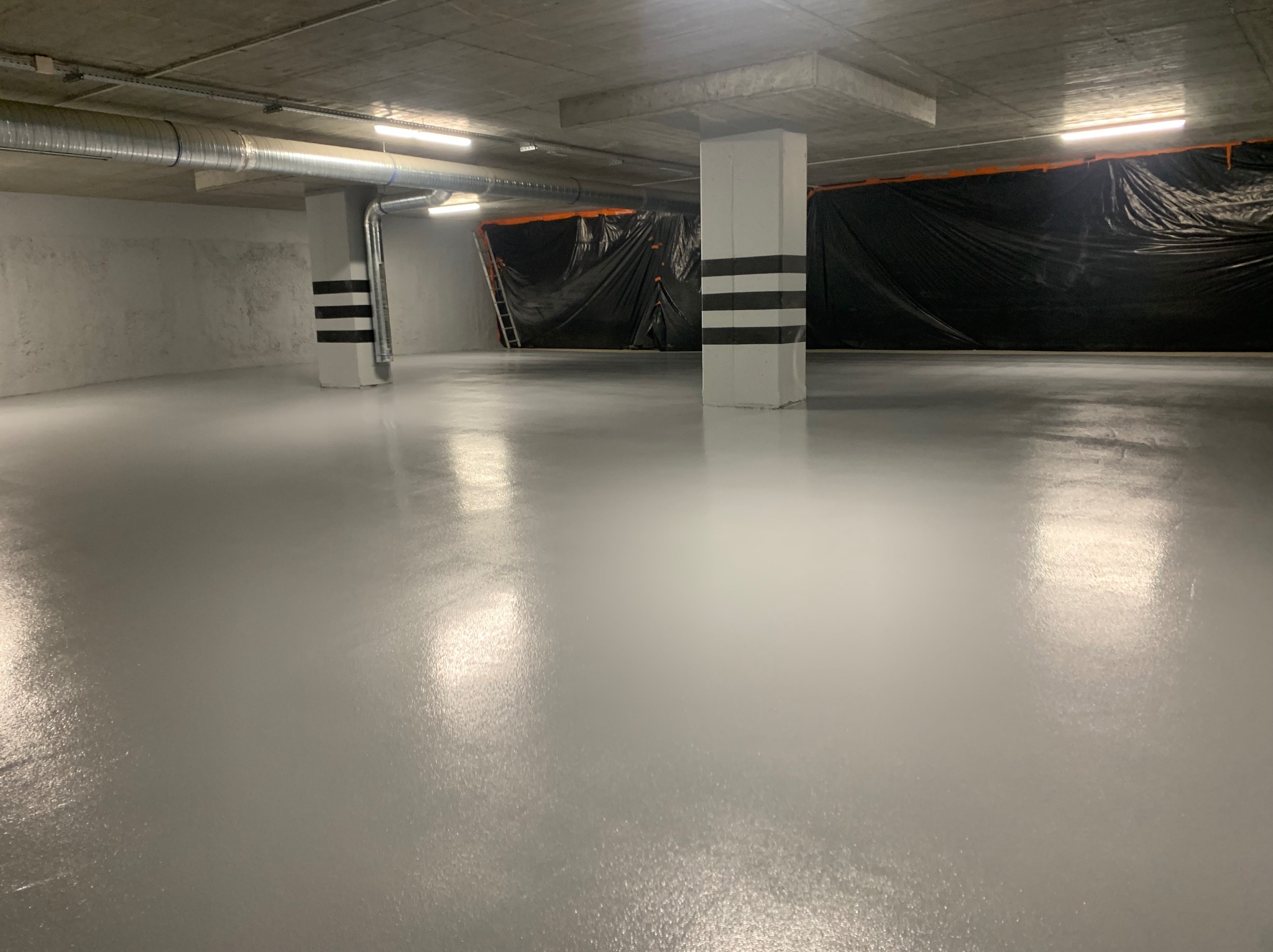 Szara, błyszcząca posadzka w hali przemysłowej, zabezpieczona folią malarską przy ścianie, z widocznymi słupami oznaczonymi czarnymi pasami.