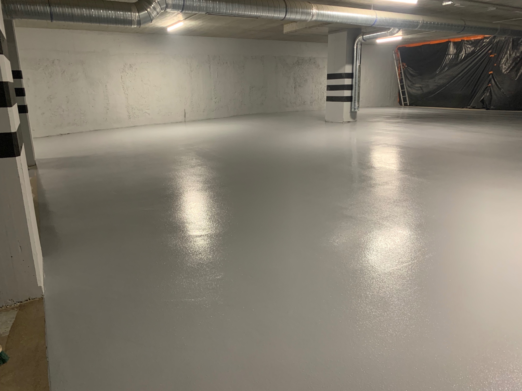 Gładka, szara posadzka w garażu podziemnym, z widocznymi rurami wentylacyjnymi pod sufitem i kolumną pomalowaną w czarno-białe pasy.