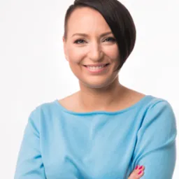 Portret uśmiechniętej kobiety z krótkimi, ciemnymi włosami, ubranej w błękitny sweter na białym tle.