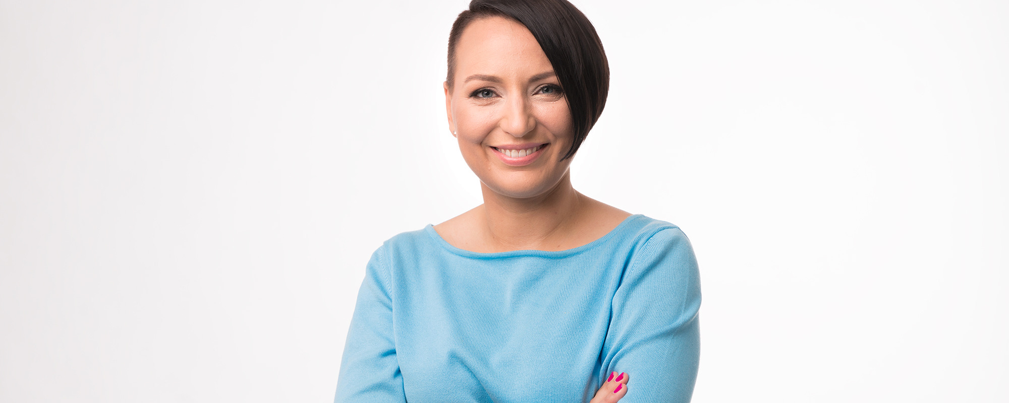 Portret uśmiechniętej kobiety z krótkimi, ciemnymi włosami, ubranej w błękitny sweter na białym tle.