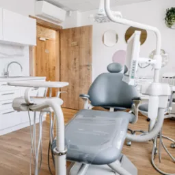 Jasne wnętrze gabinetu stomatologicznego z nowoczesnym fotelem dentystycznym w odcieniach szarości, białymi meblami z marmurowym blatem, drewnianymi drzwiami i subtelnymi okrągłymi dekoracjami...