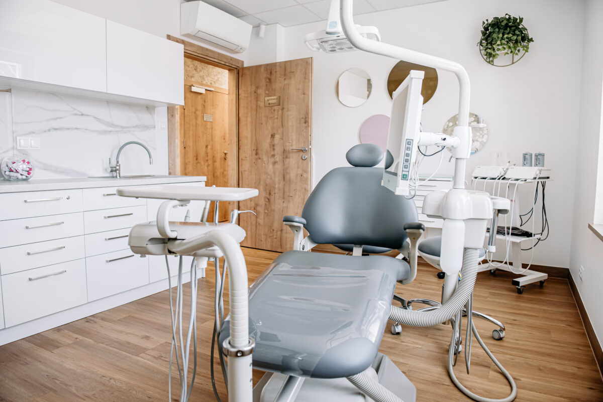 Jasne wnętrze gabinetu stomatologicznego z nowoczesnym fotelem dentystycznym w odcieniach szarości, białymi meblami z marmurowym blatem, drewnianymi drzwiami i subtelnymi okrągłymi dekoracjami...