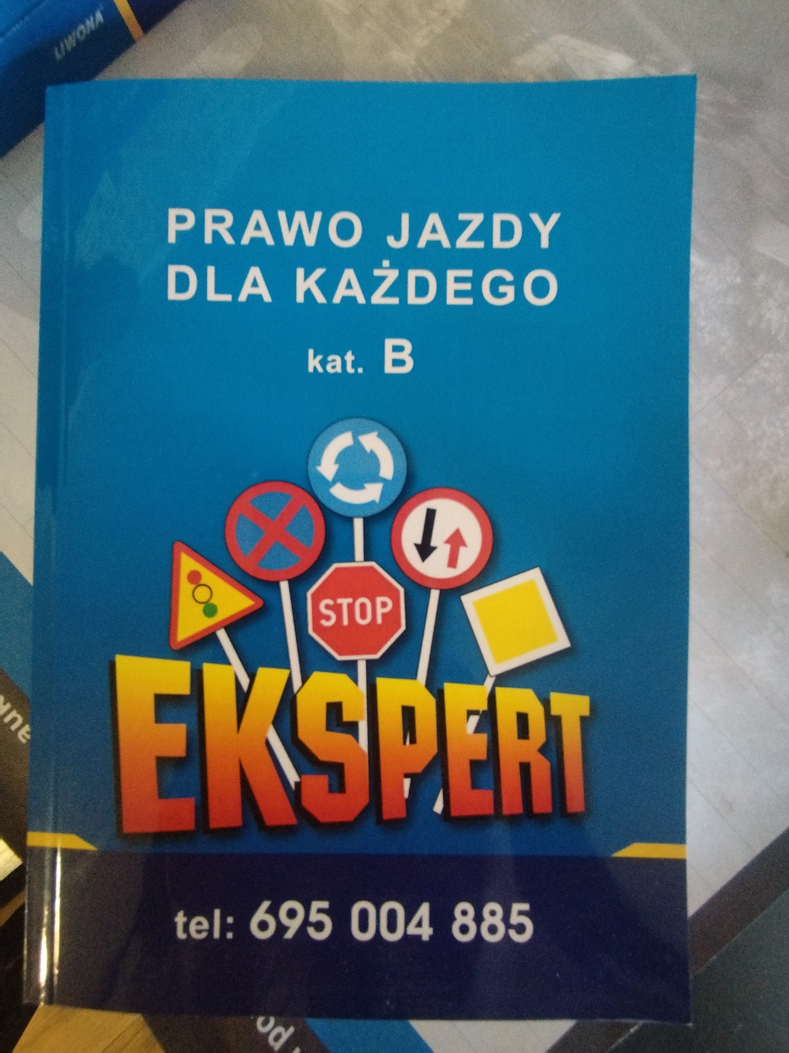 Książka z niebieską okładką 'Prawo jazdy dla każdego kat. B' z ilustracjami znaków drogowych i napisem 'EKSPERT' oraz numerem telefonu.