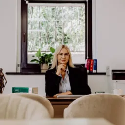 Portret kobiety prawnika w okularach, siedzącej za biurkiem w jasnym gabinecie, z figurką Temidy i książkami prawniczymi w tle.