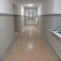 FRW STANISZEWSKI Sp. z.o.o - Długi korytarz w budynku biurowym po remoncie: świeżo pomalowane ściany z dwukolorowym wykończeniem, jasna podłoga, widoczne narzędzia malarskie i wiadro z farbą w tle.