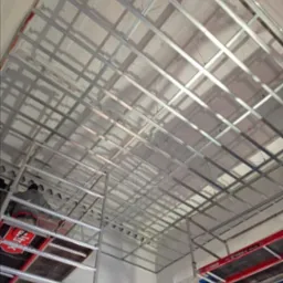 Montaż metalowej konstrukcji sufitu podwieszanego z płyt kartonowo-gipsowych, widok z dołu, fragment rusztowania z czerwoną platformą roboczą po lewej stronie.