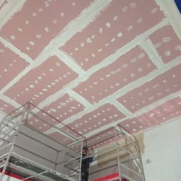 FRW STANISZEWSKI Sp. z.o.o - Montaż płyt kartonowo-gipsowych na suficie, widoczne łączenia wypełnione masą szpachlową, pracownik na rusztowaniu.