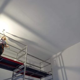FRW STANISZEWSKI Sp. z.o.o - Pracownik na rusztowaniu maluje sufit wałkiem z długą rączką, widoczne cienie na świeżo pomalowanej powierzchni.