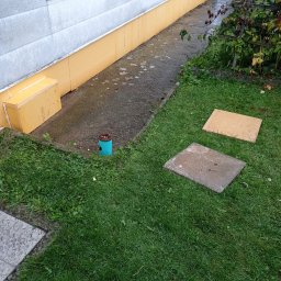 FRW STANISZEWSKI Sp. z.o.o - Ocieplenie fundamentów budynku z żółtą izolacją termiczną, widoczne fragmenty trawnika, betonowego chodnika i studzienki kanalizacyjnej.