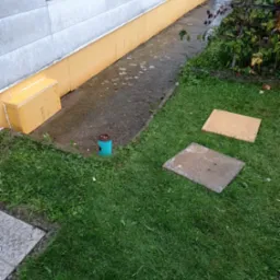 Ocieplenie fundamentów budynku z żółtą izolacją termiczną, widoczne fragmenty trawnika, betonowego chodnika i studzienki kanalizacyjnej.