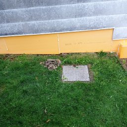 FRW STANISZEWSKI Sp. z.o.o - Ocieplenie fundamentu budynku z widoczną żółtą płytą XPS, fragmentem szarej elewacji i trawnikiem z metalową klapą rewizyjną.