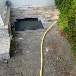 FRW STANISZEWSKI Sp. z.o.o - Wykończenie elewacji przy wejściu do budynku z dekoracyjnym kamieniem, kratką wentylacyjną i żółtym wężem ogrodowym na brukowanym chodniku.