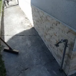 Fragment elewacji budynku wykończony dekoracyjną okładziną z kamienia naturalnego, widoczna instalacja wodna i zamiataczka z drewnianym trzonkiem, w tle pracownik w ubrudzonych spodniach roboczych.