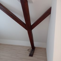 FRW STANISZEWSKI Sp. z.o.o - Wnętrze poddasza z drewnianą konstrukcją nośną w kształcie litery Y, bielone ściany i jasna podłoga, widoczne elementy instalacji elektrycznej przy podstawie słupa.